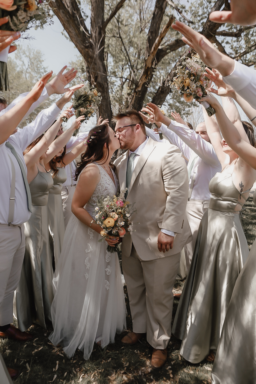 Bride and groom share a kiss under raised bouquets as their wedding party in sage green dresses and light suits celebrates around them.