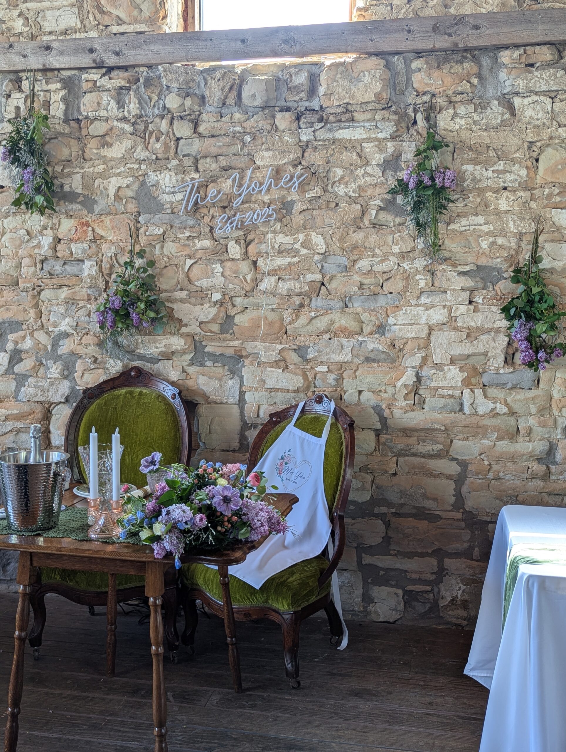 Rustic wedding sweetheart table with vintage green chairs, lilac floral arrangements, candles, champagne bucket, and a custom neon sign reading “The Yohes Est. 2025” against a stone wall backdrop.